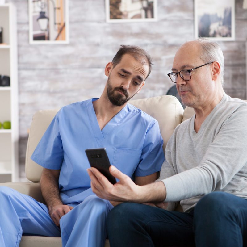 Retired man in modern nursing home sitting on couch with his health visitor. Senior man using mobile phone.
